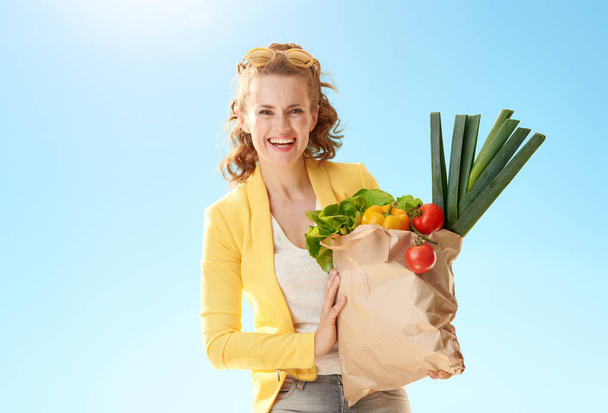 Portrait of happy stylish woman in yellow jacket with paper bag with groceries against blue sky - Fotó, kép