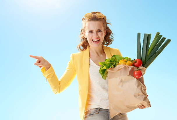 happy young woman in yellow jacket with paper bag with groceries pointing at something against blue sky - Foto, Bild