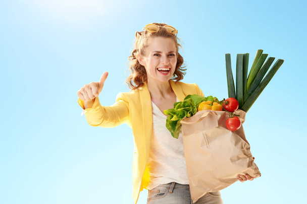 happy young woman in yellow jacket with paper bag with groceries showing thumbs up against blue sky - Foto, immagini