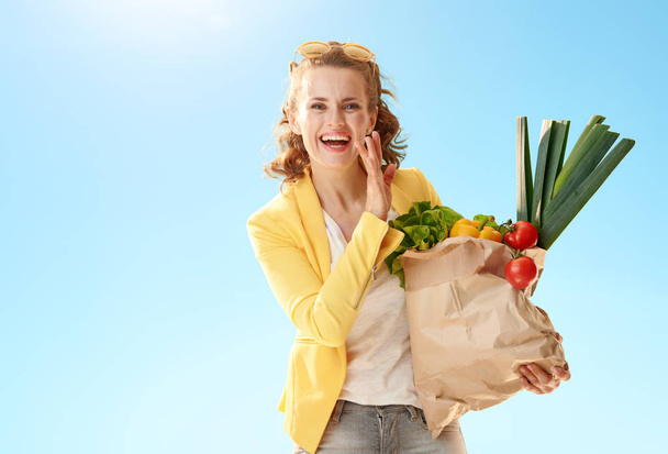 smiling modern woman in yellow jacket with paper bag with groceries telling exciting news against blue sky - Zdjęcie, obraz