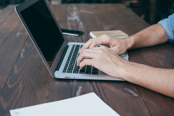 Men's hands are printed on a laptop on a wooden brown table in a cafe with free Wi-Fi, in the foreground a notebook. The concept for a freelancer works anytime, anywhere. Side view, concept - Photo, Image