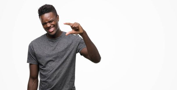 Молодой африканский американец в серой футболке улыбается и уверенно жестикулирует рукой, делая знак размера пальцами, глядя в камеру. Концепция измерения
. - Фото, изображение
