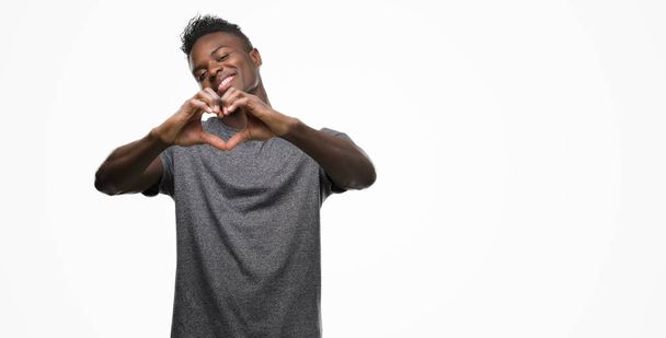 Молодой африканский американец в серой футболке, улыбающийся в любви, показывает символы сердца и форму руками. Романтическая концепция
. - Фото, изображение