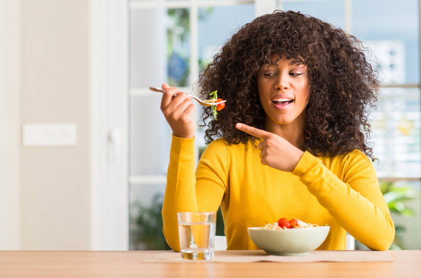 African american woman eating pasta salad at home very happy pointing with hand and finger - Photo, Image