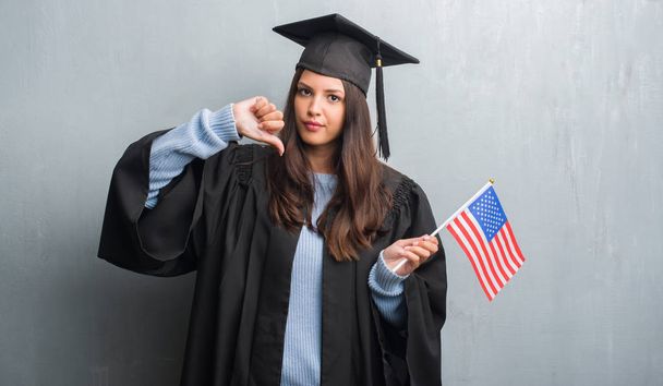 Jeune femme brune sur le mur gris grunge portant uniforme diplômé tenant drapeau de l'Amérique avec le visage en colère, signe négatif montrant aversion avec les pouces vers le bas, concept de rejet
 - Photo, image