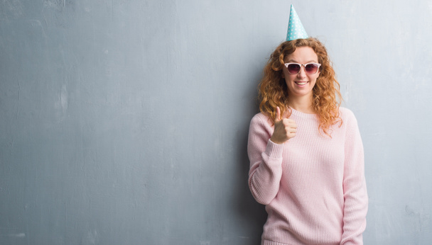 Jeune femme rousse sur un mur gris grunge portant un bonnet d'anniversaire heureux avec un grand sourire faisant signe ok, pouce levé avec les doigts, excellent signe
 - Photo, image
