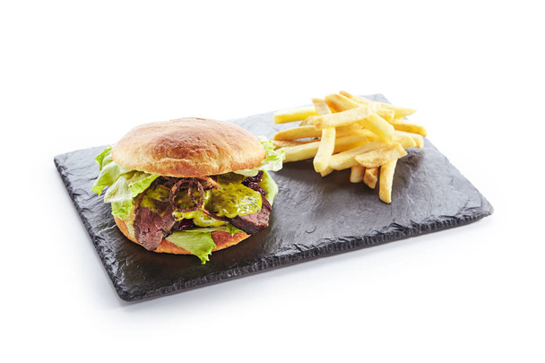 Classic Roast Beef Burger with Mustard, Fried Onions, Lettuce, Green Salsa Verde Sauce and French Fries Garnish. Beefburger with Medium Rare Steak on Natural Black Stone Plate Isolated on White - Photo, image