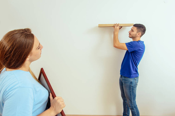 Pareja decora su nuevo hogar mientras el hombre instala estante en la pared
 - Foto, Imagen