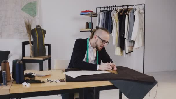 Young bearded fashion designer draw sketches using ruler, cut out cloth for future clothes. Fabric, pattern and sewing tools lie on the sewing table in front of a a tailor and young designer. Small - Metraje, vídeo
