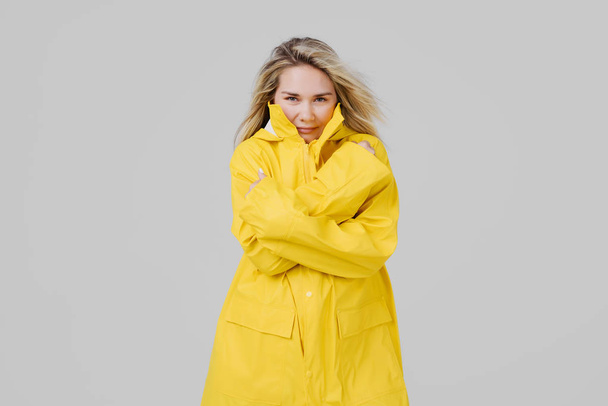 Young blonde woman in a yellow raincoat on a gray background is frozen from the rain and trying to keep warm - 写真・画像