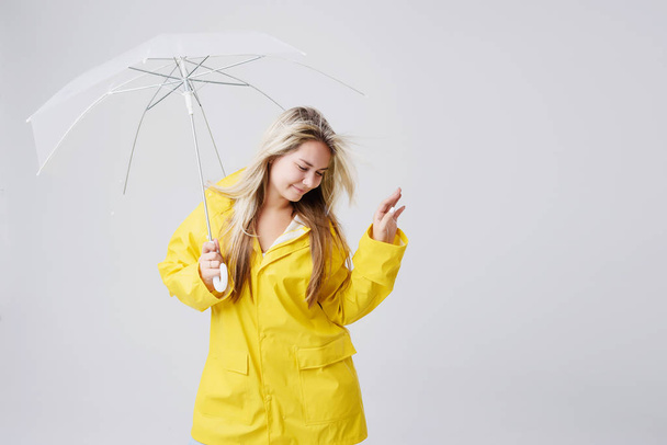 Blonde woman wearing yellow raincoat holding transparent umbrella checking weather if it is raining. Protected against rain and hurricane - Foto, afbeelding