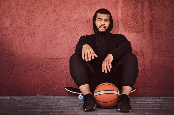 Афро-американських бородатий хлопець, одягнений в чорний балахон і спортивні шорти сидить на скейтборді з баскетболу і, спираючись на стіні. - Фото, зображення