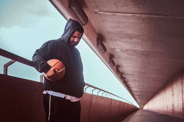 Dark-skinned bearded guy dressed in a black hoodie and sports shorts leaning on a guardrail while standing with basketball, looking down. - Photo, Image