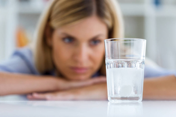 Shot of unhealthy young woman looking at a glass with an effervescent pill at home. - Foto, Imagen