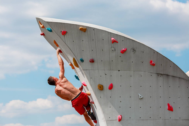 Photo from back of young sporty man in red shorts hanging on wall for rock climbing against blue sky with clouds - Foto, immagini