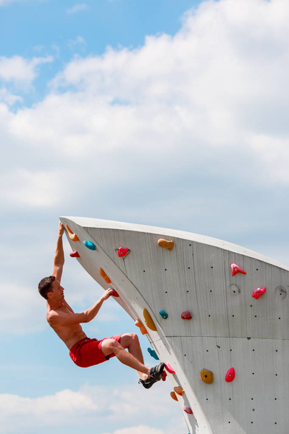 Photo of trained sports guy in red shorts practicing on wall for rock climbing against blue sky with clouds - 写真・画像