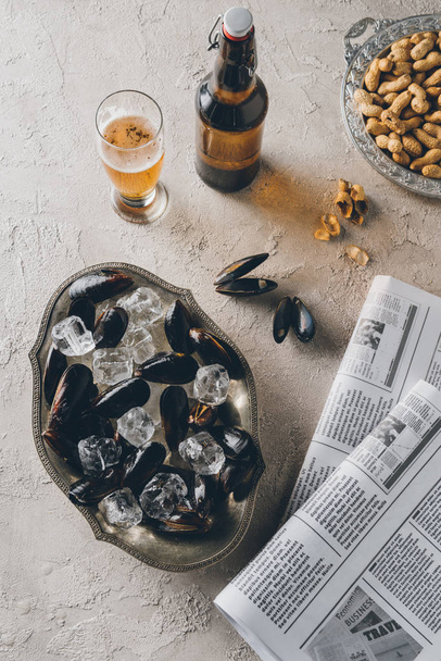 top view of mussels with ice cubes, beer, peanuts and newspapers on concrete surface - Foto, Bild