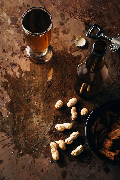 top view of arranged peanuts, baked breads, bottle and glass of beer on rust tabletop - Photo, Image