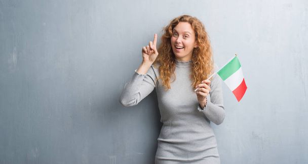 Giovane donna rossa sopra la bandiera grigia grunge wall holding d'Italia sorpresa da un'idea o domanda che punta il dito con la faccia felice, numero uno
 - Foto, immagini