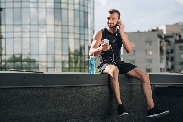 handsome cheerful sportsman listening music and using smartphone on roof  - Foto, Imagem
