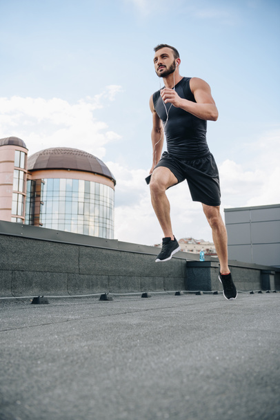 low angle view of handsome sportsman running and listening music with smartphone on roof - Foto, Imagem