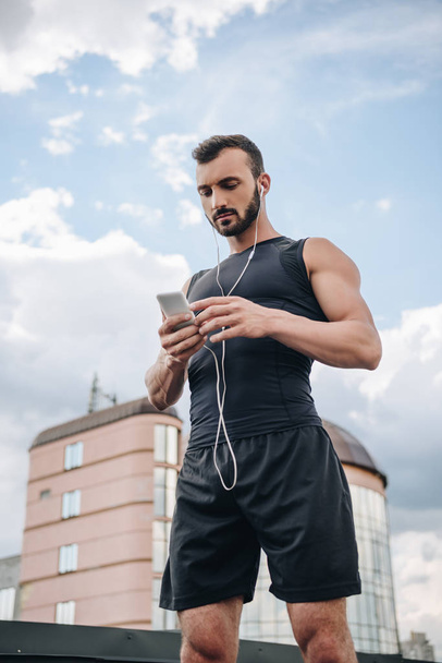 low angle view of handsome sportsman listening music and using smartphone on roof - Fotoğraf, Görsel
