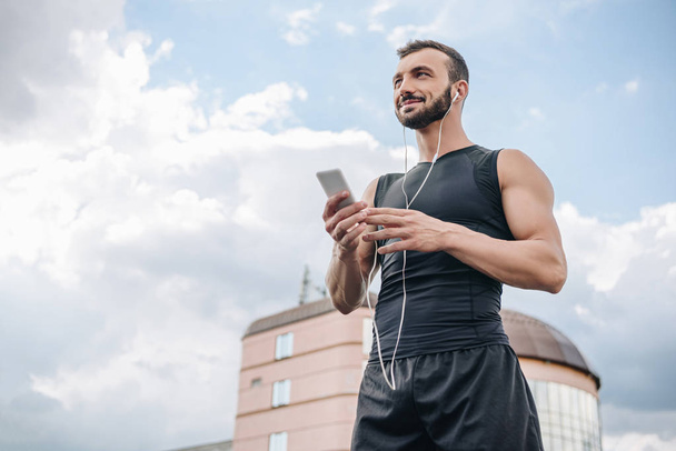 low angle view of cheerful sportsman listening music with smartphone and looking away on roof  - Foto, afbeelding