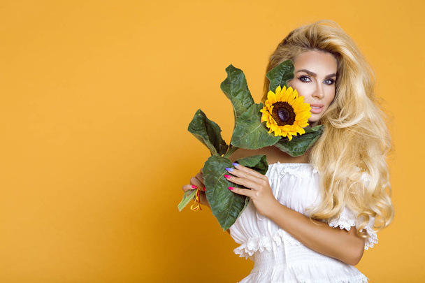 Porträt einer schönen Frau mit langen blonden Haaren, die ein weißes Kleid trägt und Sonnenblumen in der Hand hält. - Foto, Bild