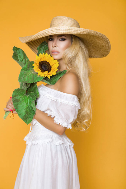 Portrait of a beautiful woman with long blond hair, wearing a white dress and hat, holding sunflowers. - Fotoğraf, Görsel