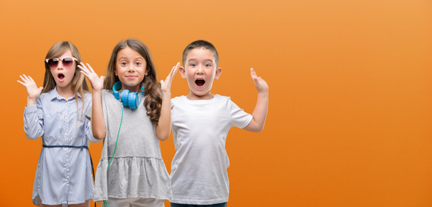 Group of boy and girls kids over orange background very happy and excited, winner expression celebrating victory screaming with big smile and raised hands - Photo, Image