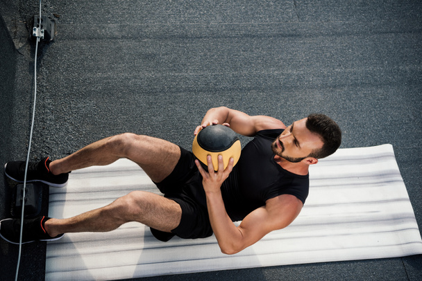 Blick aus der Vogelperspektive auf einen gut aussehenden Sportler, der Sit Ups mit Medizinball auf einer Yogamatte auf dem Dach macht - Foto, Bild