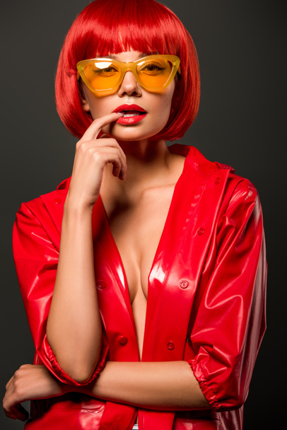 close-up portrait of beautiful young woman in red latex jacket on bare skin and vintage yellow sunglasses looking at camera isolated on grey - Photo, image