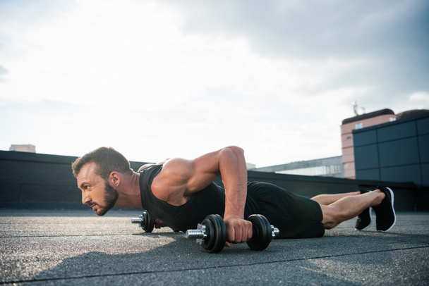 side view of handsome sportsman doing push ups with dumbbells on roof - Photo, Image