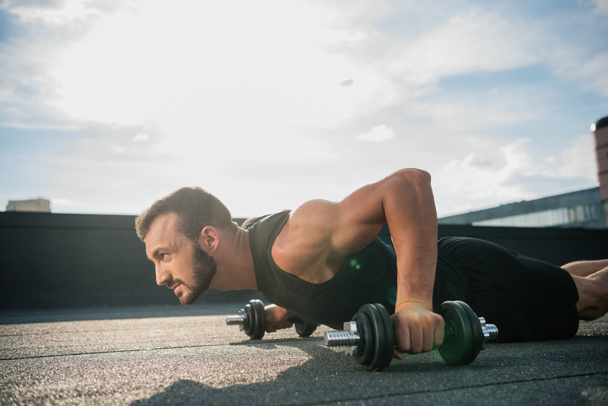 side view of handsome sportsman doing plank on dumbbells on roof  - Photo, Image