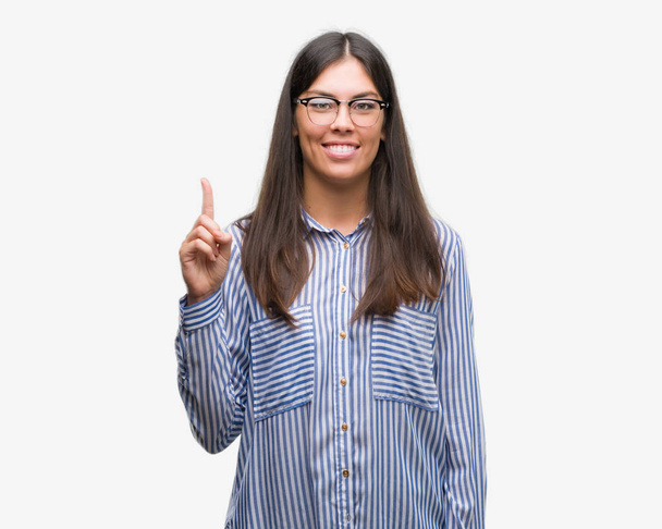 Mladé krásné Hispánská obchodní žena ukazující a směřující nahoru s prstem číslo jedna při úsměvu a spokojené. - Fotografie, Obrázek