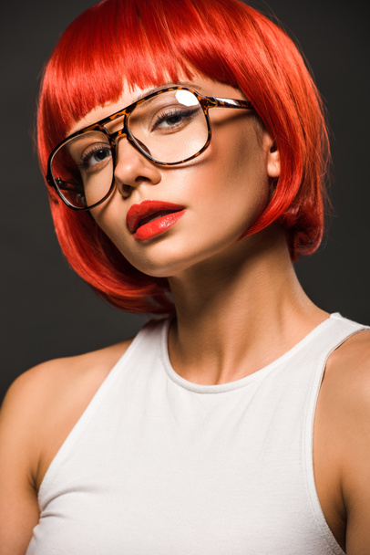 attractive young woman with red bob cut and stylish eyeglasses looking at camera isolated on grey - Fotografie, Obrázek