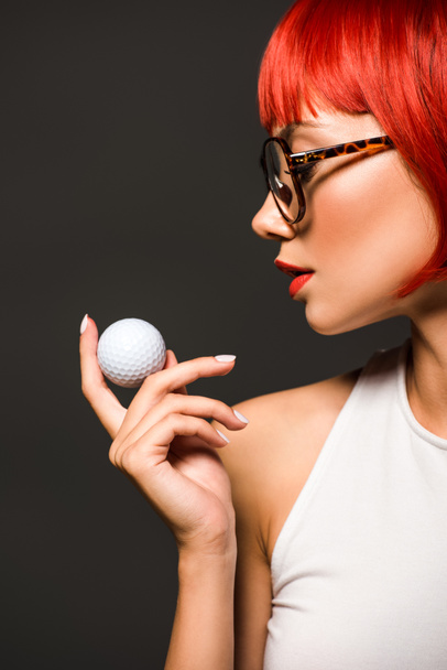 赤いボブ カットおよび灰色に分離されたゴルフ ・ ボールを保持しているスタイリッシュな眼鏡の美しい若い女性のクローズ アップの肖像画 - 写真・画像