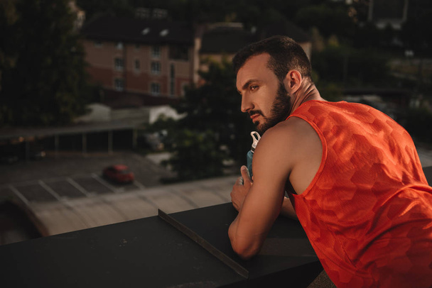 handsome sportsman holding bottle of water and leaning on railing on roof - Fotografie, Obrázek