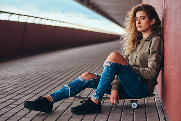 Porträt einer jungen Frau in Kapuzenpulli und zerrissener Jeans, die an einer Wand lehnt, während sie auf einem Skateboard am Brückensteg sitzt. - Foto, Bild