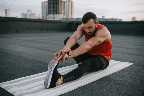 handsome sportsman stretching on yoga mat on roof - Фото, изображение