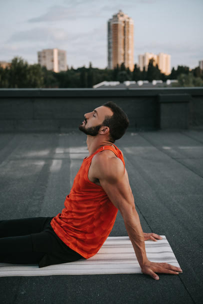 side view of handsome man sitting on yoga mat with closed eyes on roof - Photo, Image