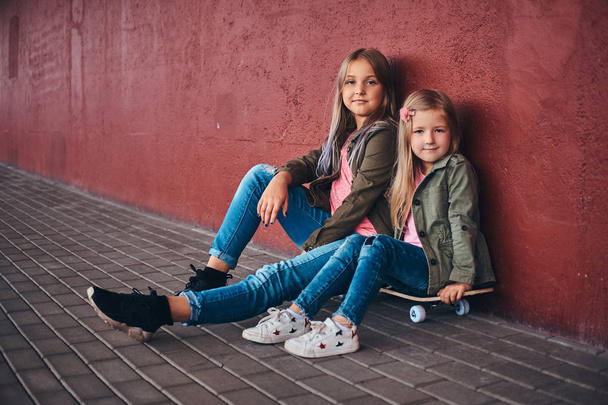 Portrait of two cute little sisters dressed in trendy clothes leaning on a wall while sitting on a skateboard at bridge footway. - Fotoğraf, Görsel