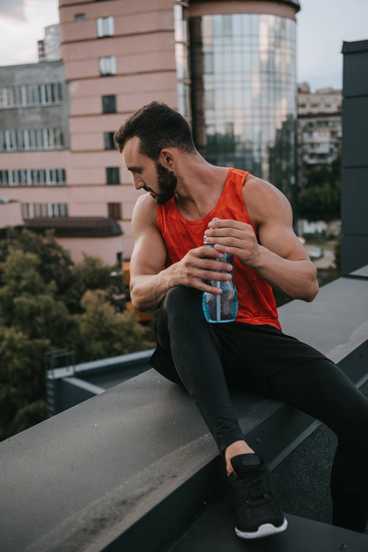 komea urheilija istuu katolla kaide pullo vettä ja katselee alas
 - Valokuva, kuva