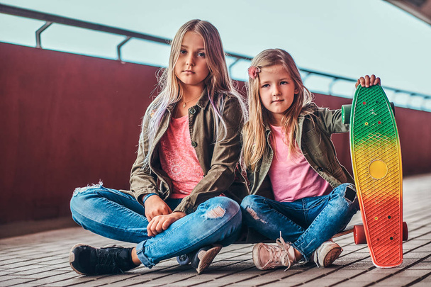 Porträt zweier kleiner Schwestern in trendigen Klamotten, die zusammen auf einem Skateboard auf einem Brückensteg sitzen. - Foto, Bild