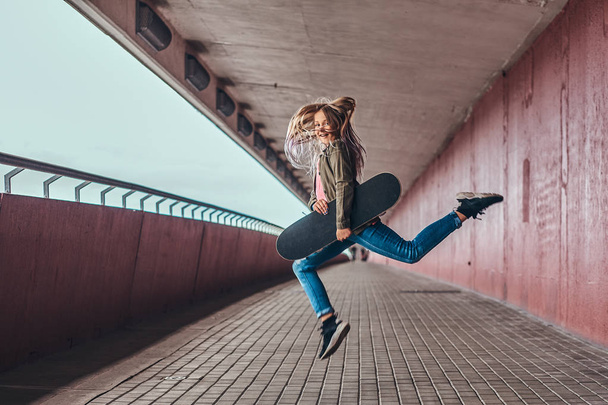 Estudante feliz com cabelo loiro vestido com roupas da moda salta com um skate no pé da ponte
. - Foto, Imagem