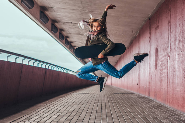 Happy schoolgirl with blonde hair dressed in trendy clothes jumps with a skateboard at bridge footway. - 写真・画像