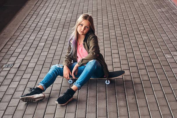 Schoolgirl with blonde hair dressed in trendy clothes sitting on a skateboard at a bridge footway. - Foto, afbeelding