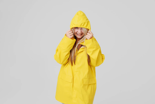 Young woman blonde in a yellow raincoat, with a hood on a head background hides from a rain on a grey background - Foto, immagini