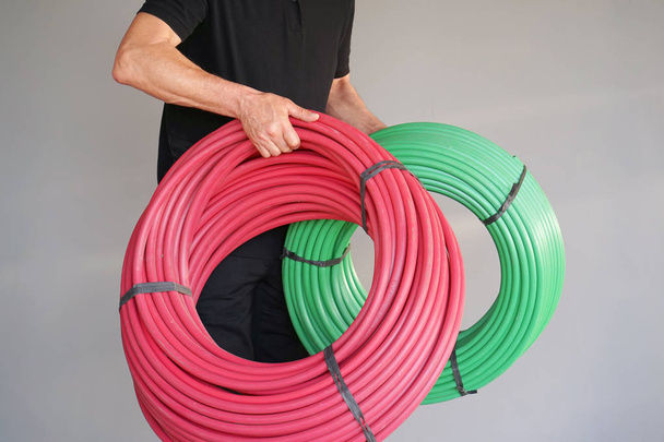 El electricista hombre sostiene verde y rojo un rollo de un tubo de protección eléctrica sobre un fondo gris. Concepto de servicios industriales y privados
. - Foto, Imagen