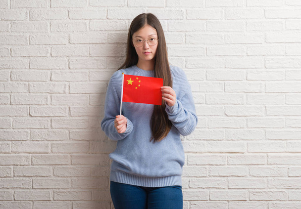 Fiatal kínai nő gazdaság Kína zászlaja, magabiztos kifejezés, a gondolkodás súlyos Szúró Szembenéz tégla falon - Fotó, kép